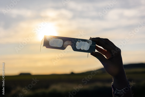 Fotografija Hand holding protective solar eclipse glasses against a setting sun, safely obse