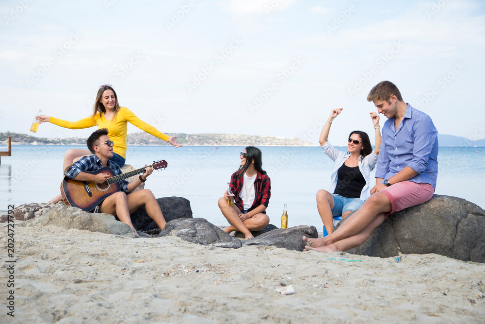 Obraz premium Friends enjoy a casual day at the beach, with one person playing an acoustic guitar while others sit and socialize, a serene coastal landscape in the background.