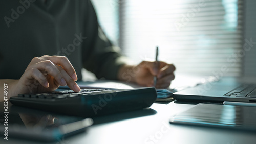 Photos Close-up of accountant calculating budget and writing report beside laptop in modern office, showing finance analysis