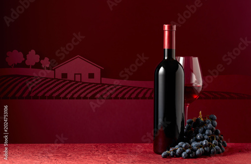 Bottle and glass of red wine with blue grapes on a red background.