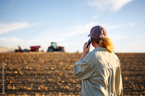 Woman farmer talking to smartphone while observing tractor sowing crop and agricultural work in cultivated field
