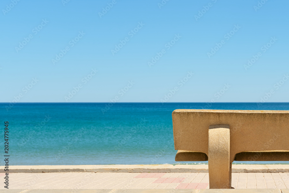 Fototapeta premium Modern concrete bench overlooking a calm Mediterranean beach and turquoise sea under clear blue sky. Minimal coastal composition with peaceful summer atmosphere and copy space.