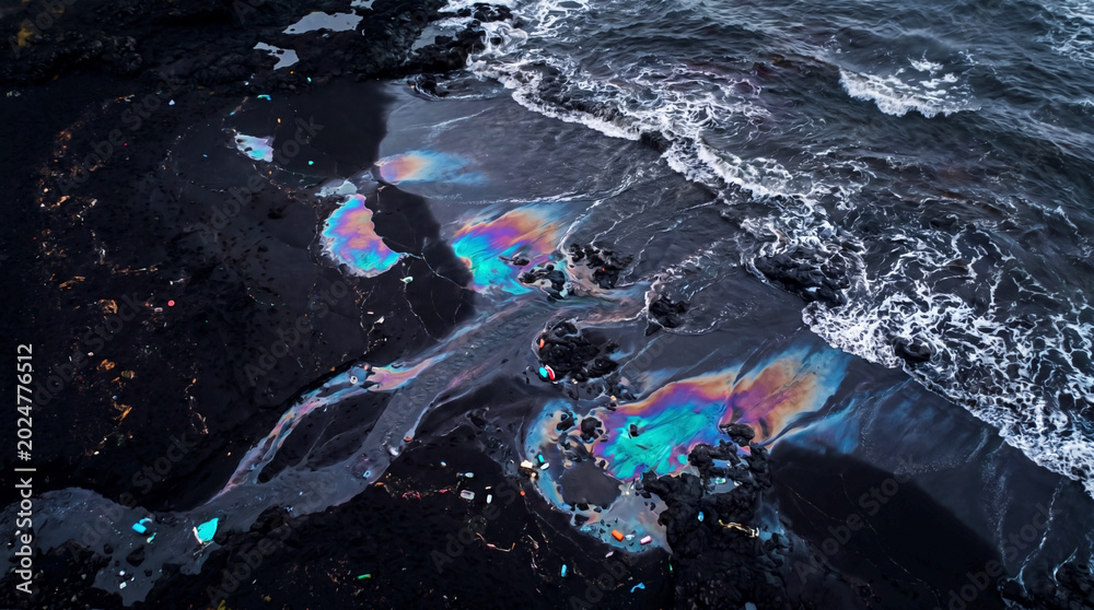 Fototapeta premium Toxic oil slick creates colorful patterns across polluted shoreline water and sand. 