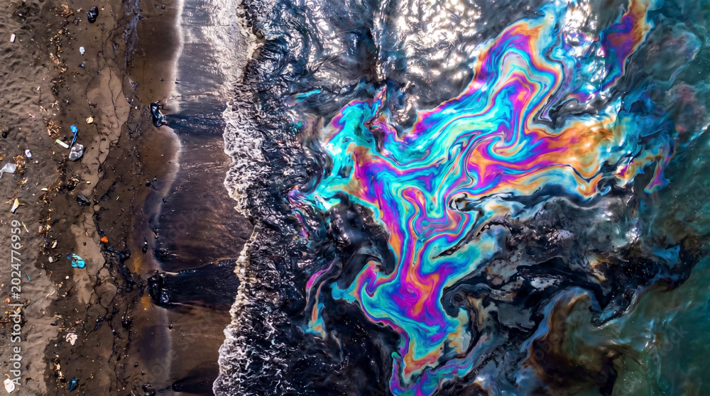 Fototapeta premium Toxic oil slick creates colorful patterns across polluted shoreline water and sand.