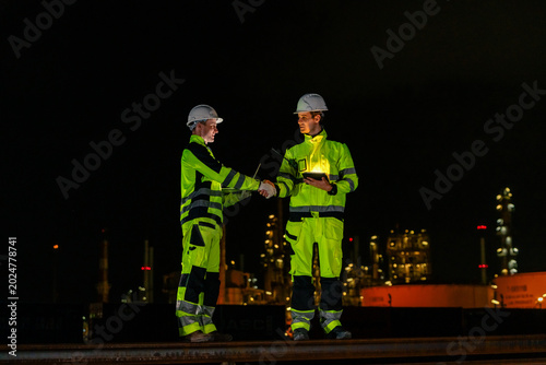 Professional male industrial engineers shaking hands at night shift illuminated petrochemical refinery plant representing successful partnership for corporate oil and gas energy business growth.