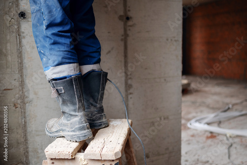 Quadro su tela A close-up of a construction worker's dusty black safety boots standing on a makeshift wooden stepladder against a bare concrete wall with copy space