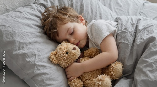Peaceful young boy peacefully sleeping on a cozy bed holding a teddy bear, serene childhood moment in a soft, gray setting, perfect for parenting, childhood, and comfort themes.
