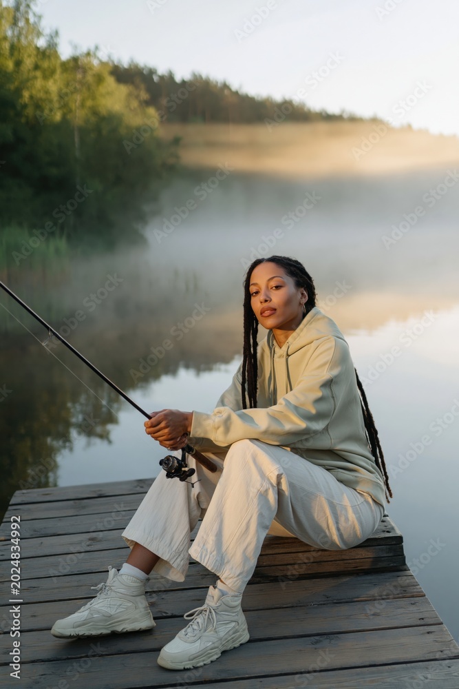 Fototapeta premium Young woman fishing on dock by misty lake at sunrise