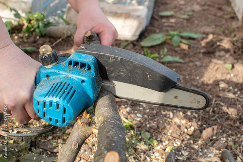 Fotografi Close-up of a person using a cordless mini chainsaw to cut dry tree branches on the ground in the backyard