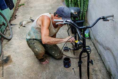 Obraz na płótnie Middle aged man restoring and painting a bicycle frame outdoors in a modest residential backyard while a cat relaxes nearby