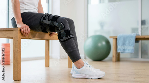 Person wearing an orthopedic knee brace for injury recovery. Patient sitting on a bench in a physical therapy clinic. Joint support and rehabilitation concept