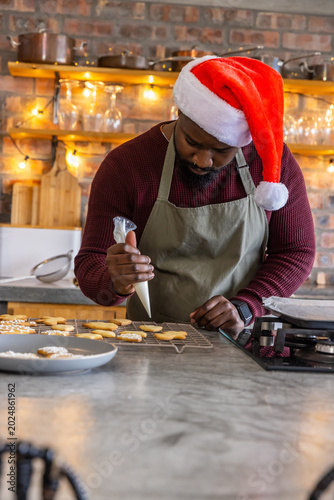 African American man decorating cookies in festive kitchen using piping bag while wearing Santa hat