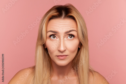 Middle aged woman looking up and examining forehead wrinkles to check skin condition and aging signs for rejuvenation treatment isolated on pink background
