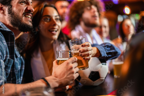 Two sports fans toasting with beer next to a soccer ball at a pub bar counter.