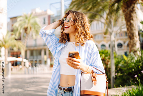 A young woman stands in a city park surrounded by palm trees. She smiles as she holds her phone, enjoying the sunny weather. The backdrop features buildings and a lively atmosphere.