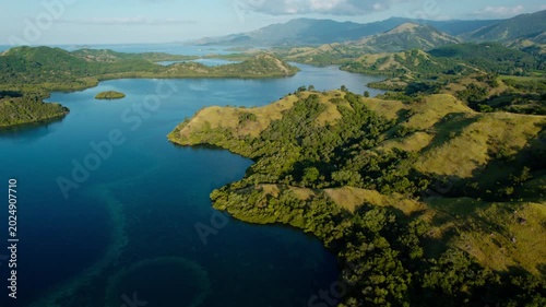 Drone shot pulling back and tilting down over green savanna hills and tropical blue sea bays in Riung 17 Islands National Park, Flores.
