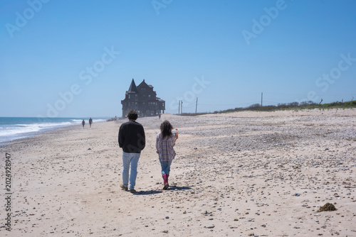Photography Bright coastal scene of a man and woman walking together along a sandy beach toward a distant oceanfront house under clear blue skies, capturing a peaceful summer seaside atmosphere