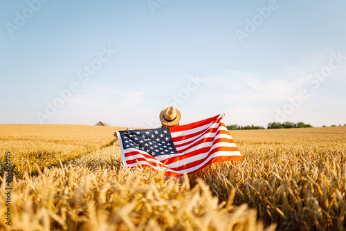 A woman stands among tall ears of wheat, waving American flags and smiling. She wears a straw hat and enjoys the sunset. The concept is celebration, freedom, and independence.