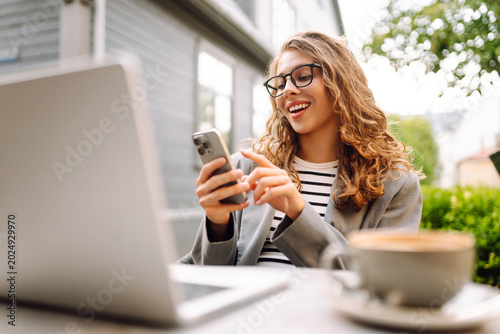 Woman sits at table outside cafe, smiling and using phone. Laptop and a cup of coffee are on table. Female freelancer works on terrace, phone in hand. Concept of remote work, technology.