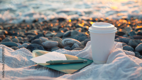 White paper coffee cup with envelope letter pen on soft blanket over pebble beach during glowing sunset by sea, concept of mental health self discovery self development emotional healing mindfulness