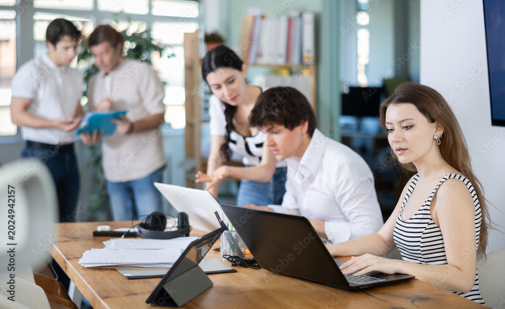 Fototapeta premium Young woman at her desk in modern office and working on laptop among group of colleagues