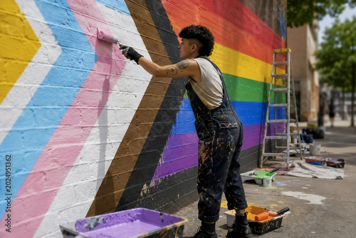 An artist wearing overalls uses a paint roller to create a large progress pride flag mural on an urban brick wall. Tools and a ladder sit on the sidewalk.