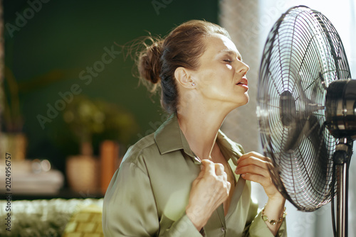A middle-aged woman, distressed by heat, leans into an electric fan, eyes closed, pulling collar for relief. Sunlight highlights her face, emphasizing intense discomfort of a summer heatwave indoors.