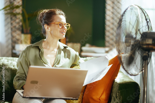 An adult woman with orange glasses smiles, eyes closed, enjoying the cool breeze from a fan. On a green sofa with laptop & papers, she embodies comfort & productivity in a warm home office setting