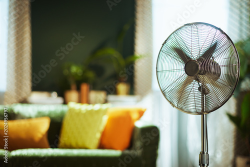 A modern stand fan with spinning blades is prominently featured, providing refreshing air circulation in a comfortable living room with a green velvet sofa and natural light, ideal for heat relief.