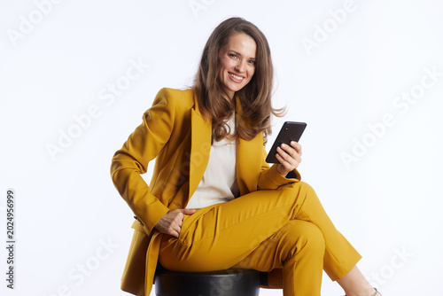 Foto A smiling executive in a mustard yellow pantsuit holds a smartphone, looking directly at the camera