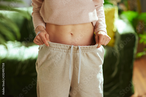 A woman in a pink crop top demonstrates her fitness progress by pulling at the waistband of her oversized grey sweatpants in a bright living room.