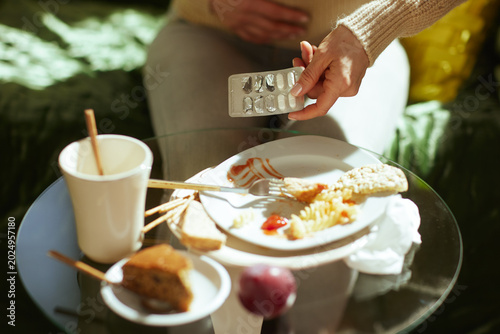 Tableau sur toile A woman rests one hand on her stomach while holding a pack of pills, surrounded by leftover pasta and cake, suggesting indigestion from unhealthy food habits