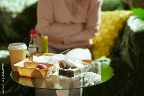 Sitting on a couch with arms crossed over her stomach, a woman experiences post-meal distress near a table of sushi and coffee, highlighting digestive issues.