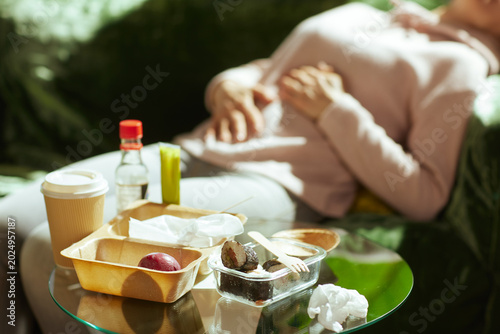Lying on a sofa, a woman rests her hands on her bloated stomach after eating takeout. Sunlight filters through as she recovers from the discomfort of a heavy, unhealthy meal.