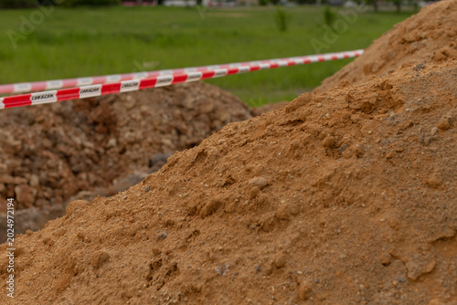 Playground sand delivery near open trench, safety tape outlines work zone, loose granular material sitting on grassy field, nearby apartment blocks and cloudy