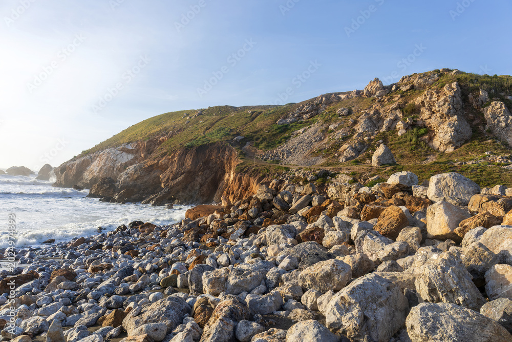 Fototapeta premium A rugged coastal landscape at Rockaway Beach in Pacifica, California, featuring a rocky shoreline scattered with large boulders and surf-worn stones beneath steep ocean cliffs.
