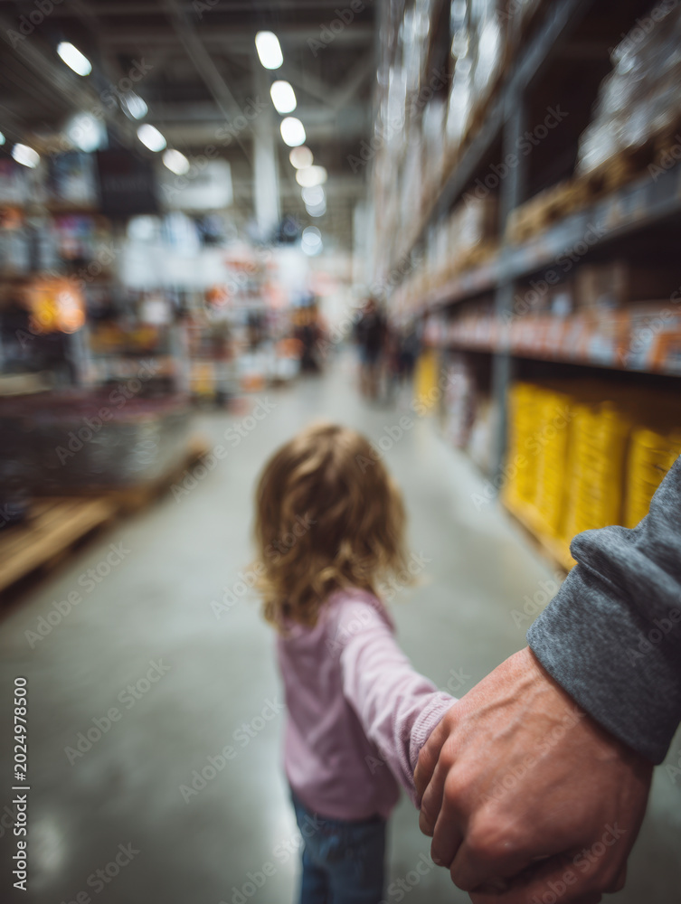 Fototapeta premium Parent holding child's hand while walking through a large busy warehouse-style store aisle filled with shelves stocked with various merchandise and supplies