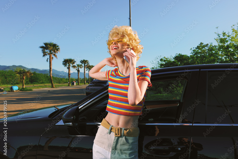 Fototapeta premium Smiling woman with curly blonde hair leaning beside a black car in a sunny outdoor scene, wearing a rainbow striped top and light shorts, conveying carefree retro summer lifestyle.