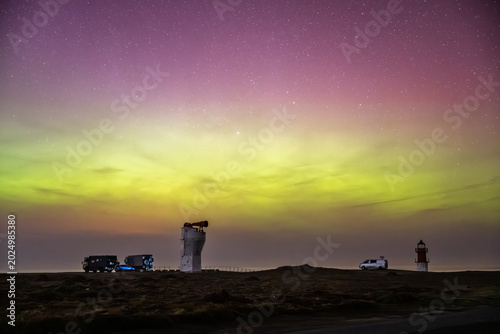 The historic white foghorn tower and the red-and-white Winkie Lighthouse stand under a vast night sky painted with green and pink Northern Lights,Point of Ayre, Isle of Man.