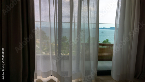 Soft morning light filtering through sheer curtains with a serene ocean view from a modern hotel room balcony, creating a calm tropical vacation atmosphere with minimalist interior aesthetics.