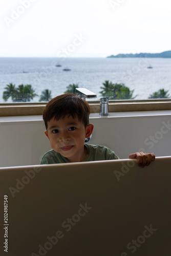close up photo of a boy in the bathtub area looking at the camera with a background of a sea and island view with several coconut trees