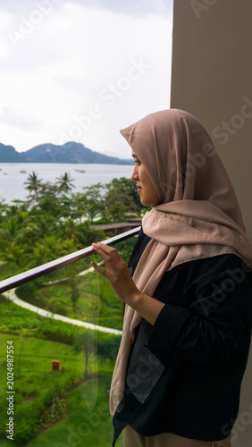 A vertical photo of a beautiful Asian girl wearing a hijab enjoying the beautiful view from a hotel balcony in Indonesia. A moment during a summer vacation.