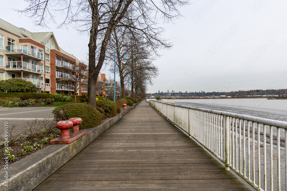 Fototapeta premium Quiet Waterfront Boardwalk Along River With Residential Buildings, Trees, and Railings in New Westminster
