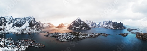 Atmospheric drone panorama of Reine and nearby islands in the Lofoten archipelago, northern Norway. Golden hour light reflects across Arctic fjords between snowy mountains and remote fishing villages.