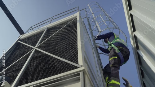 Engineer maintenance hvac cooling tower ladder safety harness technician industrial worker inspects metal structure under blue sky during service check rooftop climb to maintain and monitor