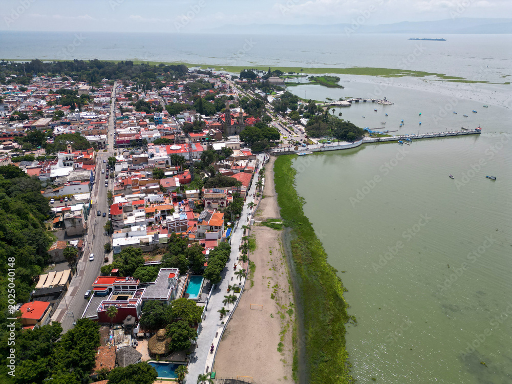 Fototapeta premium High-angle drone perspective showcases the colorful architecture and bustling malecon of Chapala, Mexico, bordering the calm, green waters of Lake Chapala under a bright sky.