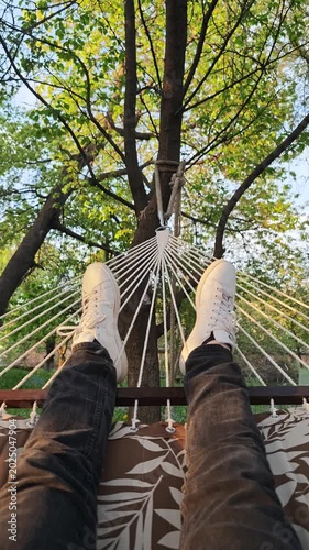 A serene first-person perspective shot of someone unwinding in a comfortable white rope hammock, featuring their feet clad in stylish white sneakers and dark jeans, as they slowly sway amidst lush