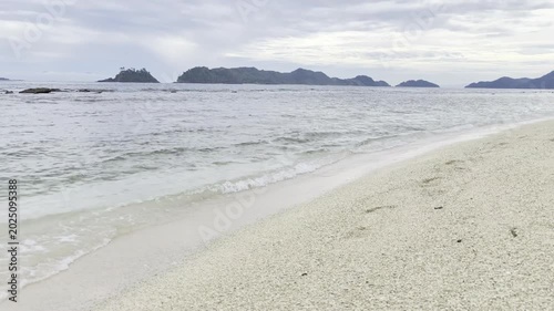 Footprints in the pristine sand leading towards the gentle waves of a serene ocean, with picturesque islands gracing the horizon under an overcast sky