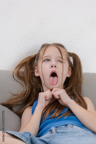 Young playful girl with pigtail making silly expression face tongue out feeling bored sitting on grey couch wearing blue shirt white background casual childhood home environment