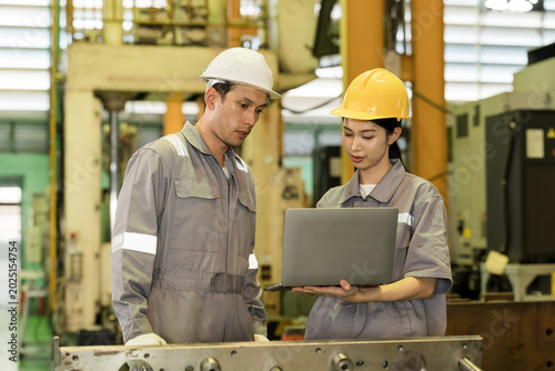Asian adult female engineer presents laptop data to caucasian adult male colleague, reviewing component status, workflow issue and production detail beside heavy equipment in factory workshop.